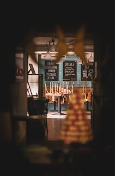 A dimly lit restaurant interior with chairs placed upside down on tables, creating a closed or after-hours atmosphere. Menu boards on the wall list drinks and specials, and pendant lights hang from the ceiling.