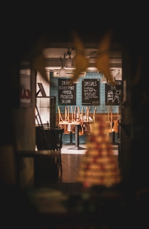 A dimly lit restaurant interior with chairs placed upside down on tables, creating a closed or after-hours atmosphere. Menu boards on the wall list drinks and specials, and pendant lights hang from the ceiling.