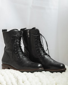A pair of black leather lace-up boots are placed on a soft, textured white surface. The boots have intricate brogue detailing on the toe and sides, with a smooth finish and sturdy soles.
