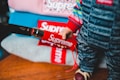 A close-up of a person wearing clothing with a bold red and white 'Supreme' logo. The attire includes a colorful, striped top and pants featuring the brand name in a repeated pattern. Several folded items, also displaying the logo, are stacked in the background.