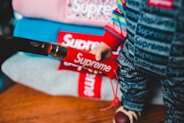 A close-up of a person wearing clothing with a bold red and white 'Supreme' logo. The attire includes a colorful, striped top and pants featuring the brand name in a repeated pattern. Several folded items, also displaying the logo, are stacked in the background.