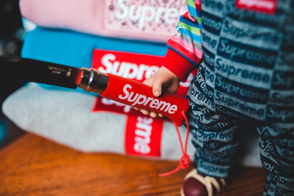 A close-up of a person wearing clothing with a bold red and white 'Supreme' logo. The attire includes a colorful, striped top and pants featuring the brand name in a repeated pattern. Several folded items, also displaying the logo, are stacked in the background.