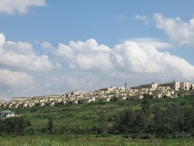 A large residential complex of uniform beige buildings stretches across a grassy hillside. The area is surrounded by lush greenery with bushes and trees in the foreground. Above, the sky is filled with fluffy white clouds scattered against a light blue background.