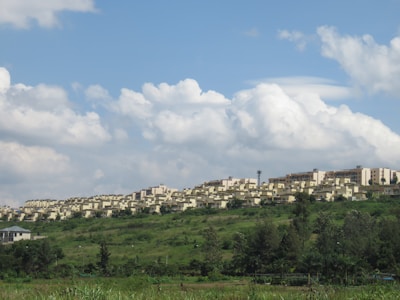 A large residential complex of uniform beige buildings stretches across a grassy hillside. The area is surrounded by lush greenery with bushes and trees in the foreground. Above, the sky is filled with fluffy white clouds scattered against a light blue background.