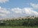 A large residential complex of uniform beige buildings stretches across a grassy hillside. The area is surrounded by lush greenery with bushes and trees in the foreground. Above, the sky is filled with fluffy white clouds scattered against a light blue background.