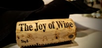A close-up view of a wine cork lying on a table. The cork has 'The Joy of Wine' printed on it in bold letters. The background consists of a blurred black napkin and a part of a wine glass.