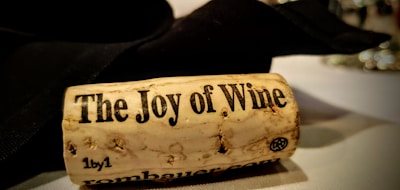 A close-up view of a wine cork lying on a table. The cork has 'The Joy of Wine' printed on it in bold letters. The background consists of a blurred black napkin and a part of a wine glass.