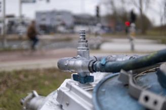A close-up view of a metallic valve connected to several thick cables, with a blurred urban background. The focus is on the industrial component in the foreground, while figures and vehicles are indistinct in the distance.