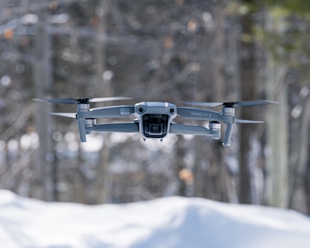 A drone hovers in the air against a blurred forest background. The foreground features snow, suggesting a cold environment. The drone's details are clear, showcasing its camera and rotors in motion.