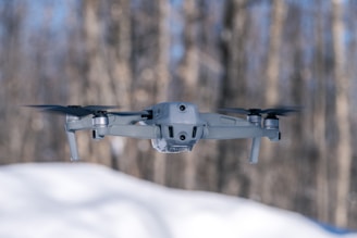 A modern drone with multiple rotors hovers in the air against a blurred background of trees. The focus is sharp on the drone, emphasizing its sleek and compact design.