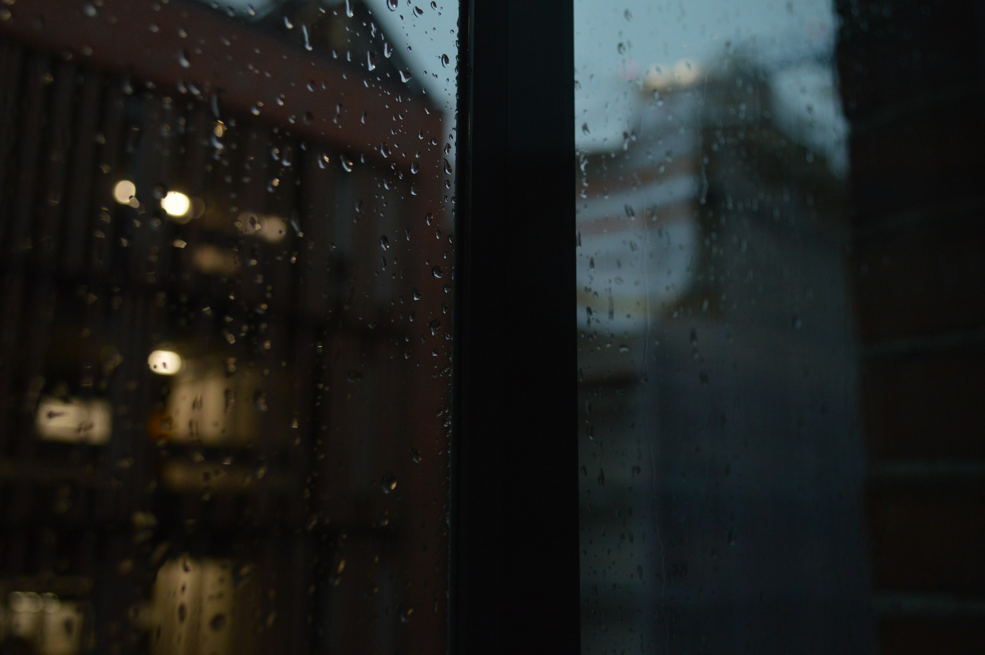 A close-up of raindrops sliding down a window, with faint silhouettes of Japanese city buildings glowing softly in the background.