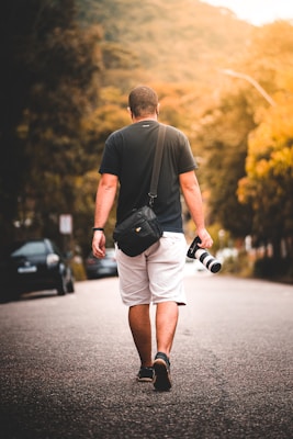 A person walks down a quiet street holding a camera with a large lens. They are wearing a black t-shirt, white shorts, and carrying a black bag. The background consists of blurred trees and sunlight, giving a warm, golden atmosphere.