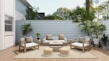 A cozy outdoor patio features modern wooden furniture with white cushions and patterned pillows. The area includes a sofa, two armchairs, a coffee table, and two round woven poufs. Lush green plants are placed around the space, along the light gray wooden fence, with a large white house wall on one side. The wooden decking and neutral rug enhance the serene atmosphere, while natural light filters through, highlighting the greenery and creating a tranquil space.