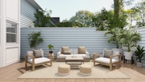 A cozy outdoor patio features modern wooden furniture with white cushions and patterned pillows. The area includes a sofa, two armchairs, a coffee table, and two round woven poufs. Lush green plants are placed around the space, along the light gray wooden fence, with a large white house wall on one side. The wooden decking and neutral rug enhance the serene atmosphere, while natural light filters through, highlighting the greenery and creating a tranquil space.