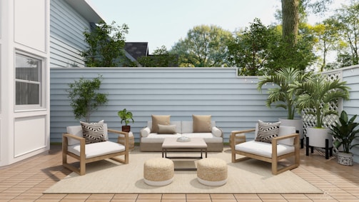 A cozy outdoor patio features modern wooden furniture with white cushions and patterned pillows. The area includes a sofa, two armchairs, a coffee table, and two round woven poufs. Lush green plants are placed around the space, along the light gray wooden fence, with a large white house wall on one side. The wooden decking and neutral rug enhance the serene atmosphere, while natural light filters through, highlighting the greenery and creating a tranquil space.