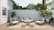A cozy outdoor patio features modern wooden furniture with white cushions and patterned pillows. The area includes a sofa, two armchairs, a coffee table, and two round woven poufs. Lush green plants are placed around the space, along the light gray wooden fence, with a large white house wall on one side. The wooden decking and neutral rug enhance the serene atmosphere, while natural light filters through, highlighting the greenery and creating a tranquil space.