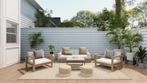 A cozy outdoor patio features modern wooden furniture with white cushions and patterned pillows. The area includes a sofa, two armchairs, a coffee table, and two round woven poufs. Lush green plants are placed around the space, along the light gray wooden fence, with a large white house wall on one side. The wooden decking and neutral rug enhance the serene atmosphere, while natural light filters through, highlighting the greenery and creating a tranquil space.