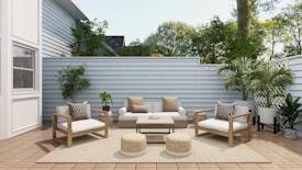 A cozy outdoor patio features modern wooden furniture with white cushions and patterned pillows. The area includes a sofa, two armchairs, a coffee table, and two round woven poufs. Lush green plants are placed around the space, along the light gray wooden fence, with a large white house wall on one side. The wooden decking and neutral rug enhance the serene atmosphere, while natural light filters through, highlighting the greenery and creating a tranquil space.
