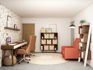 A cozy and modern home office space with a warm color palette. The room features a wooden desk with drawers and a brown swivel chair. A laptop is placed on the desk, accompanied by task lighting. To the right, there is a large orange armchair next to a tall shelf filled with books and decorative items. On the wall above the desk, there is a shelf with a clock and framed pictures. The opposite wall displays a symmetrical shelving unit with woven baskets. The floor is carpeted in gray with a circular beige rug in the center. The walls are decorated with colorful patterned wallpaper on one side.