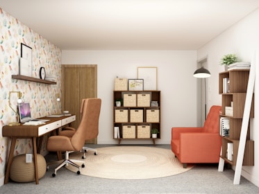 A cozy and modern home office space with a warm color palette. The room features a wooden desk with drawers and a brown swivel chair. A laptop is placed on the desk, accompanied by task lighting. To the right, there is a large orange armchair next to a tall shelf filled with books and decorative items. On the wall above the desk, there is a shelf with a clock and framed pictures. The opposite wall displays a symmetrical shelving unit with woven baskets. The floor is carpeted in gray with a circular beige rug in the center. The walls are decorated with colorful patterned wallpaper on one side.