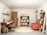 A cozy and modern home office space with a warm color palette. The room features a wooden desk with drawers and a brown swivel chair. A laptop is placed on the desk, accompanied by task lighting. To the right, there is a large orange armchair next to a tall shelf filled with books and decorative items. On the wall above the desk, there is a shelf with a clock and framed pictures. The opposite wall displays a symmetrical shelving unit with woven baskets. The floor is carpeted in gray with a circular beige rug in the center. The walls are decorated with colorful patterned wallpaper on one side.