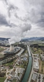 An aerial view of an industrial landscape with a large cooling tower emitting steam, surrounded by a river and several buildings including residential areas and factories. The sky is overcast with clouds, and there are mountains visible in the background.