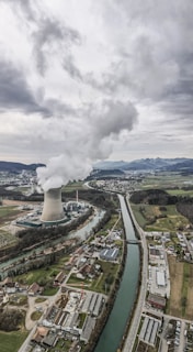 An aerial view of an industrial landscape with a large cooling tower emitting steam, surrounded by a river and several buildings including residential areas and factories. The sky is overcast with clouds, and there are mountains visible in the background.