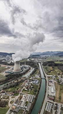 An aerial view of an industrial landscape with a large cooling tower emitting steam, surrounded by a river and several buildings including residential areas and factories. The sky is overcast with clouds, and there are mountains visible in the background.