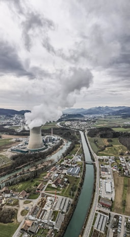 An aerial view of an industrial landscape with a large cooling tower emitting steam, surrounded by a river and several buildings including residential areas and factories. The sky is overcast with clouds, and there are mountains visible in the background.