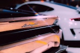 The image focuses on the rear end of a luxury sports car, prominently featuring the Lamborghini logo. Another sleek, modern car is visible in the background.
