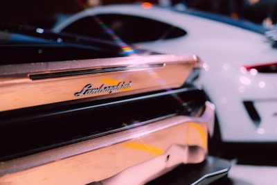 The image focuses on the rear end of a luxury sports car, prominently featuring the Lamborghini logo. Another sleek, modern car is visible in the background.
