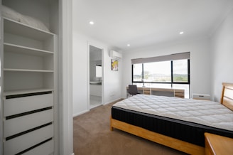 A clean, modern bedroom featuring a neatly made bed with a light-colored quilt. The room includes a large window with a scenic view, a workstation with a chair, and an air conditioning unit above. Adjacent is an open wardrobe with shelves and a doorway leading to an en suite bathroom.