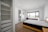 A clean, modern bedroom featuring a neatly made bed with a light-colored quilt. The room includes a large window with a scenic view, a workstation with a chair, and an air conditioning unit above. Adjacent is an open wardrobe with shelves and a doorway leading to an en suite bathroom.
