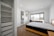 A clean, modern bedroom featuring a neatly made bed with a light-colored quilt. The room includes a large window with a scenic view, a workstation with a chair, and an air conditioning unit above. Adjacent is an open wardrobe with shelves and a doorway leading to an en suite bathroom.