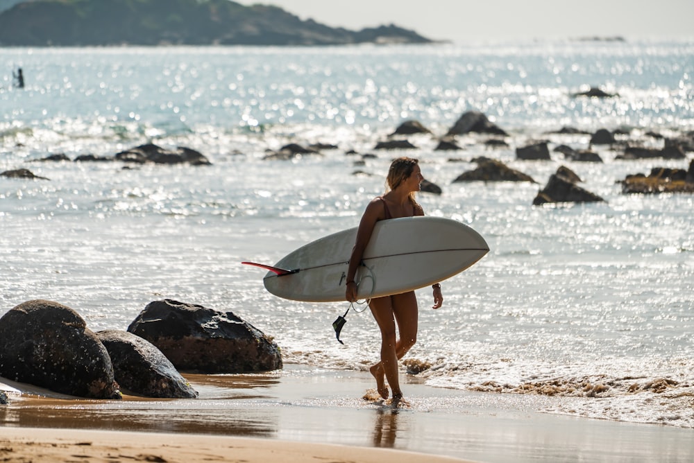 Mujer en bikini negro sosteniendo tabla de surf blanca caminando en la orilla del mar durante el día