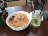 A bowl of spicy noodle soup with rich, red broth is placed on a wooden table. The soup is garnished with pieces of meat, fried tofu skin, and leafy herbs. Beside the bowl, there is a refreshing cocktail with ice, a slice of lime, and a lychee, also featuring a straw. The background shows part of a dining area with woven chairs.