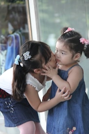 Two young children are interacting affectionately. The older child is giving a gentle kiss on the cheek to the younger child. Both are wearing denim outfits with hair accessories, standing in what seems to be a casual setting next to a large window or glass surface.