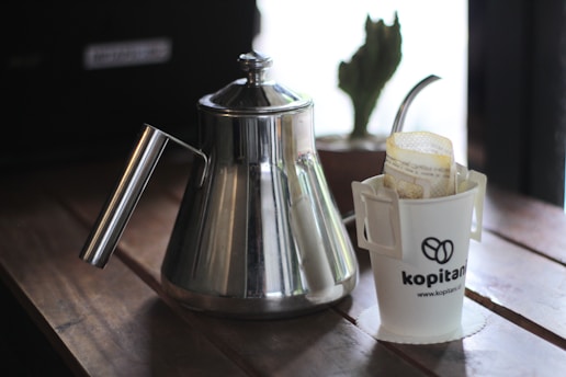 A stainless steel kettle is placed on a wooden surface beside a white cup holding a filter and branded with the logo 'kopitan'. A cactus in a small pot is partially visible in the background.