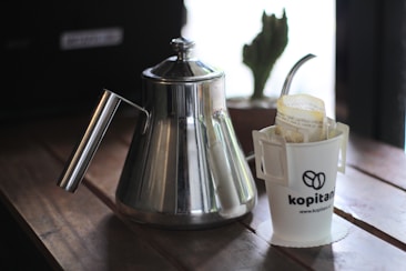 A stainless steel kettle is placed on a wooden surface beside a white cup holding a filter and branded with the logo 'kopitan'. A cactus in a small pot is partially visible in the background.