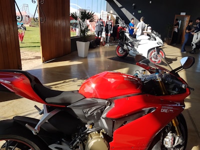 A vibrant red motorcycle is parked indoors on a polished floor. Behind it, a white motorcycle is displayed alongside several motorcycle suits on mannequins. Several people are engaged in conversation in the background. Tall wooden doors are open to a green lawn outside.