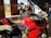 A vibrant red motorcycle is parked indoors on a polished floor. Behind it, a white motorcycle is displayed alongside several motorcycle suits on mannequins. Several people are engaged in conversation in the background. Tall wooden doors are open to a green lawn outside.