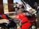 A vibrant red motorcycle is parked indoors on a polished floor. Behind it, a white motorcycle is displayed alongside several motorcycle suits on mannequins. Several people are engaged in conversation in the background. Tall wooden doors are open to a green lawn outside.