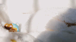 A small, colorful figurine of a bear lies against a soft, white fluffy background. The bear has a blue and brown patchwork pattern with closed eyes, as if it's sleeping. The lighting creates soft shadows and highlights on the fabric, emphasizing its texture.