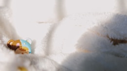 A small, colorful figurine of a bear lies against a soft, white fluffy background. The bear has a blue and brown patchwork pattern with closed eyes, as if it's sleeping. The lighting creates soft shadows and highlights on the fabric, emphasizing its texture.
