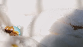 A small, colorful figurine of a bear lies against a soft, white fluffy background. The bear has a blue and brown patchwork pattern with closed eyes, as if it's sleeping. The lighting creates soft shadows and highlights on the fabric, emphasizing its texture.