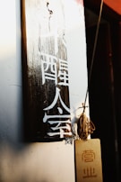 A wooden plaque with white Chinese characters hangs on a wall. The lighting is warm, casting shadows and highlighting the texture of the wood. Next to it, a small board with additional characters hangs by a string, contributing to a rustic and cultural aesthetic.