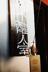 A wooden plaque with white Chinese characters hangs on a wall. The lighting is warm, casting shadows and highlighting the texture of the wood. Next to it, a small board with additional characters hangs by a string, contributing to a rustic and cultural aesthetic.