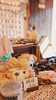 A cozy shop display featuring plush toys, with one prominently wearing a beige bow. Surrounding items include accessories and decorative pieces, with signs displaying prices. The warm lighting and lace tablecloth add a quaint and inviting atmosphere.