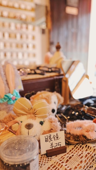 A cozy shop display featuring plush toys, with one prominently wearing a beige bow. Surrounding items include accessories and decorative pieces, with signs displaying prices. The warm lighting and lace tablecloth add a quaint and inviting atmosphere.