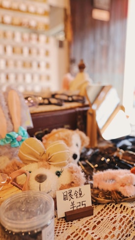 A cozy shop display featuring plush toys, with one prominently wearing a beige bow. Surrounding items include accessories and decorative pieces, with signs displaying prices. The warm lighting and lace tablecloth add a quaint and inviting atmosphere.